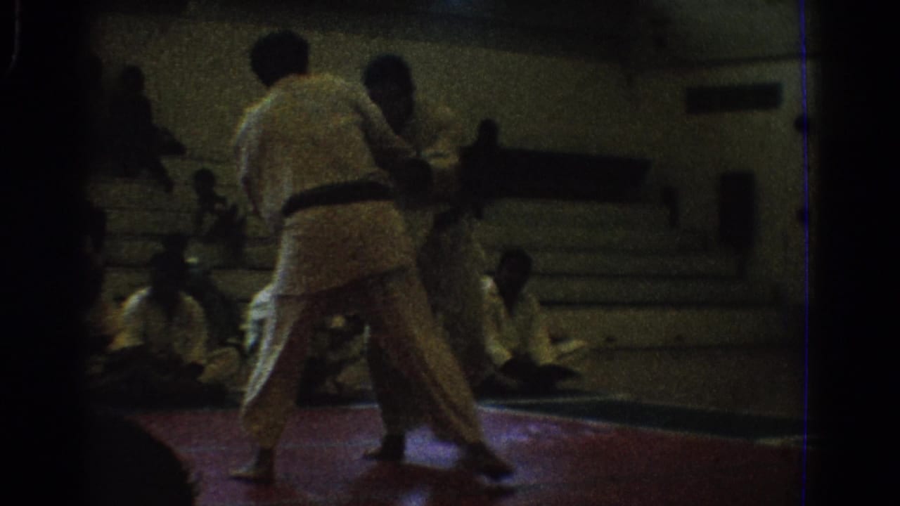 Two martial artists in white gi continue a match on a red mat in a 1966 West Covina gymnasium setting.