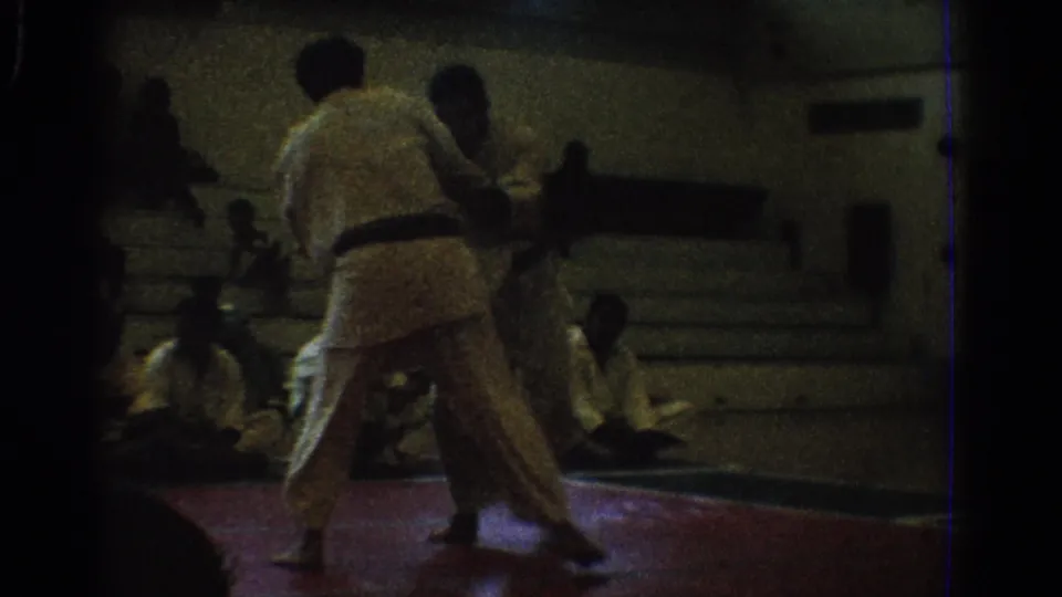 Two martial artists in white gi continue a match on a red mat in a 1966 West Covina gymnasium setting.