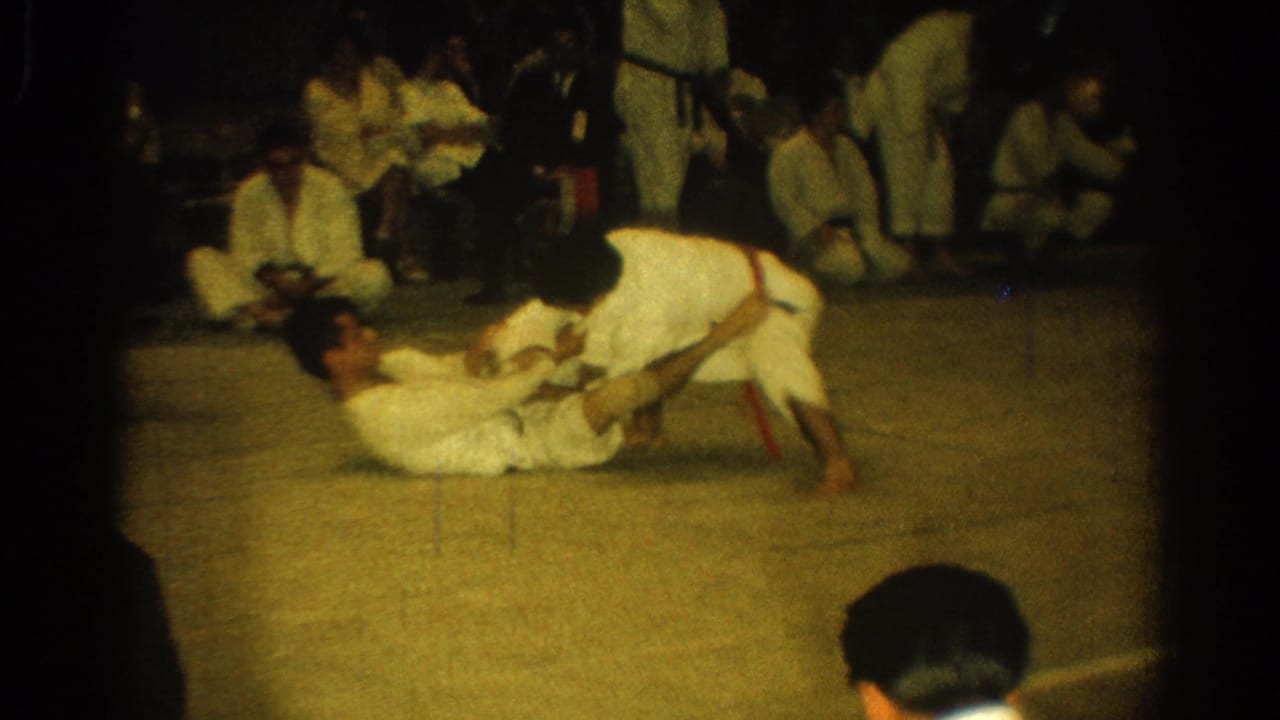 Two men in white gi grapple on a mat while others sit nearby watching.