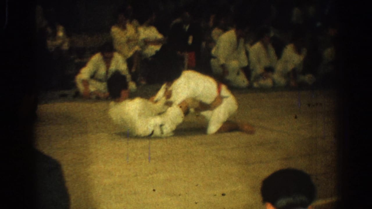 Two men in white gi continue a ground-fighting exchange on a mat, with other martial arts participants watching nearby.