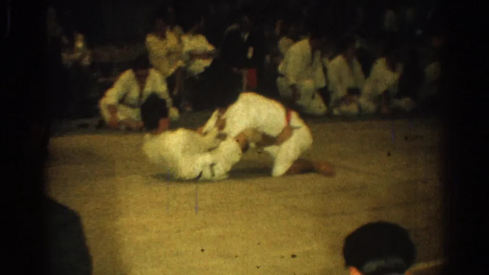 Two men in white gi continue a ground-fighting exchange on a mat, with other martial arts participants watching nearby.