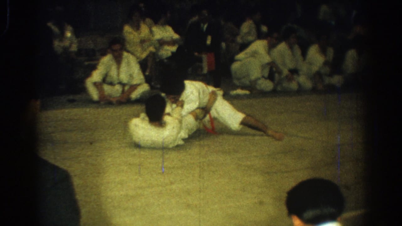 A gi-clad competitor lies on the mat while another leans across him, framed by seated observers in the background.