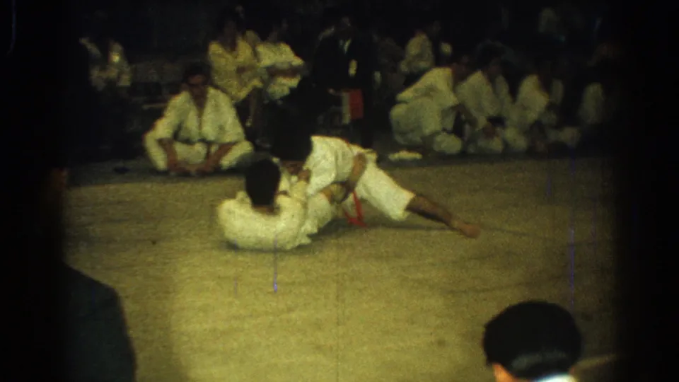 A gi-clad competitor lies on the mat while another leans across him, framed by seated observers in the background.