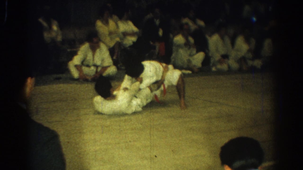 Two judo competitors grapple on a mat while spectators sit along the far side.