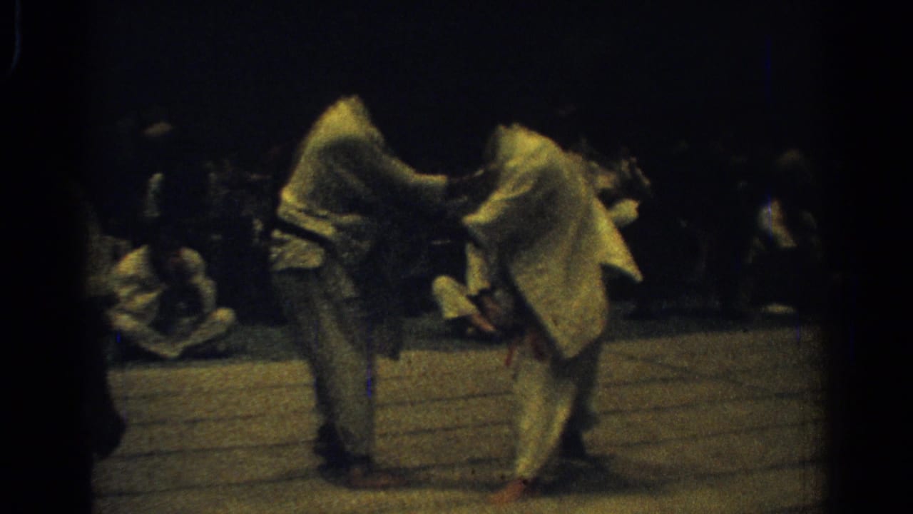 The handheld 8mm frame shows two judo competitors locked in close contact, with spectators seated around the mat.