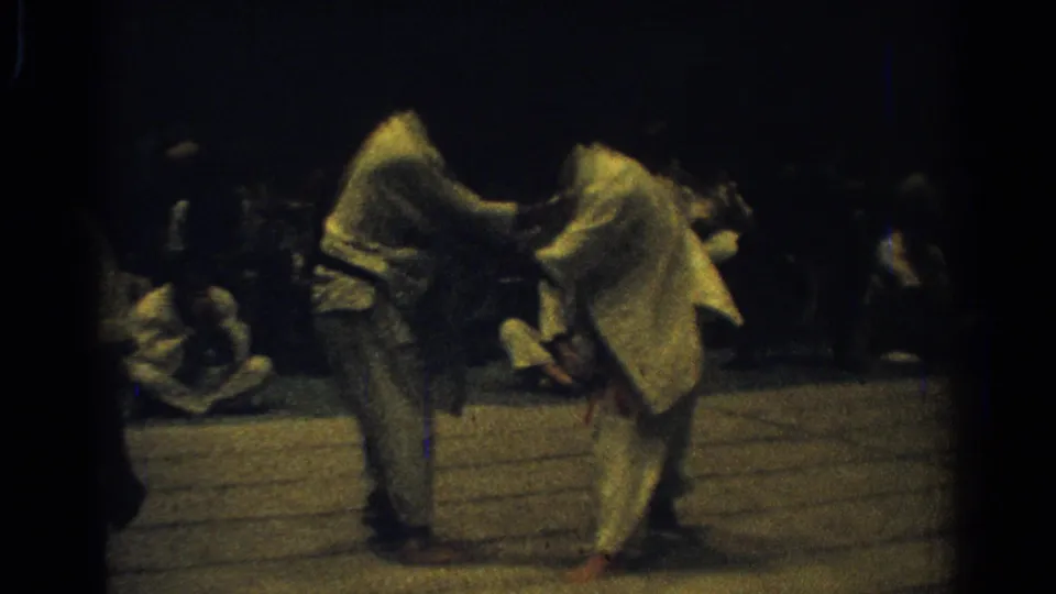 The handheld 8mm frame shows two judo competitors locked in close contact, with spectators seated around the mat.