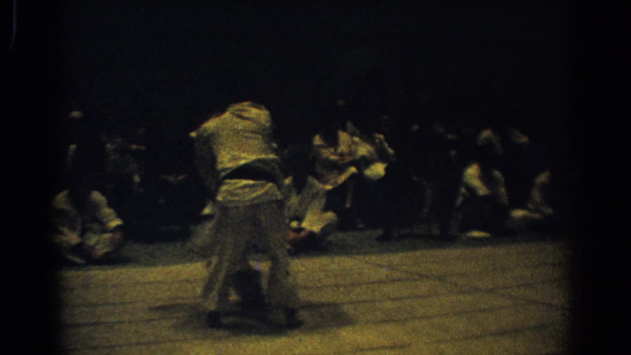 A judo competitor bends forward near another participant on the mat while spectators watch from the side of the West Covina venue.