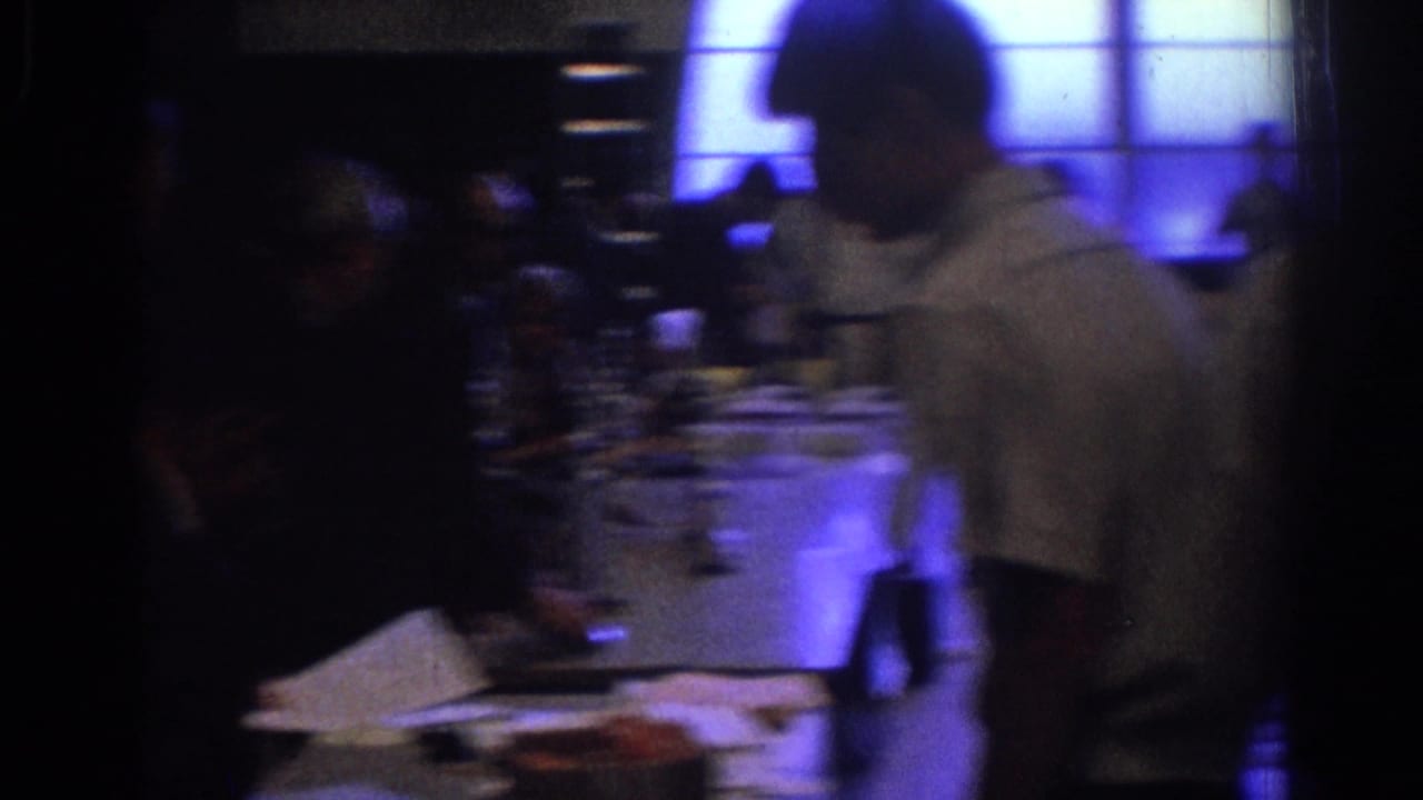 1966 West Covina home-movie frame showing a dim business or conference interior with blurred attendees, tabletop papers, and blue window light.