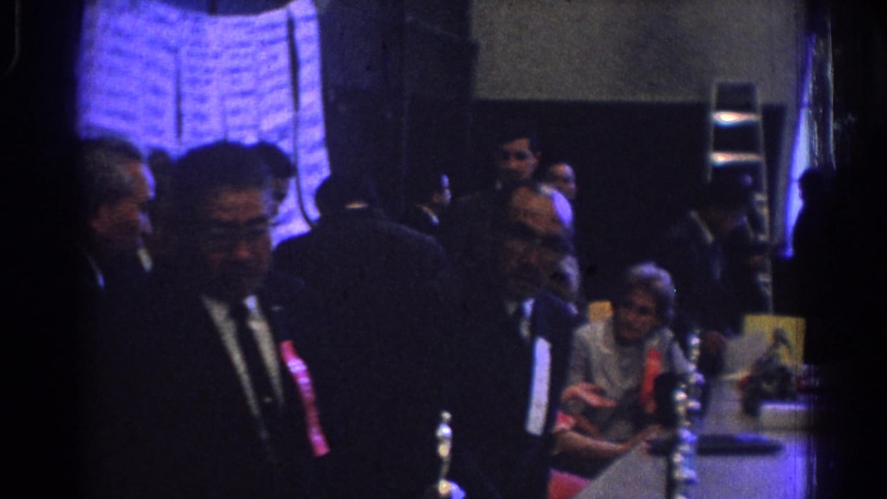 1966 West Covina home-movie frame showing suited attendees near tables with papers and bottles, with written charts along the wall.
