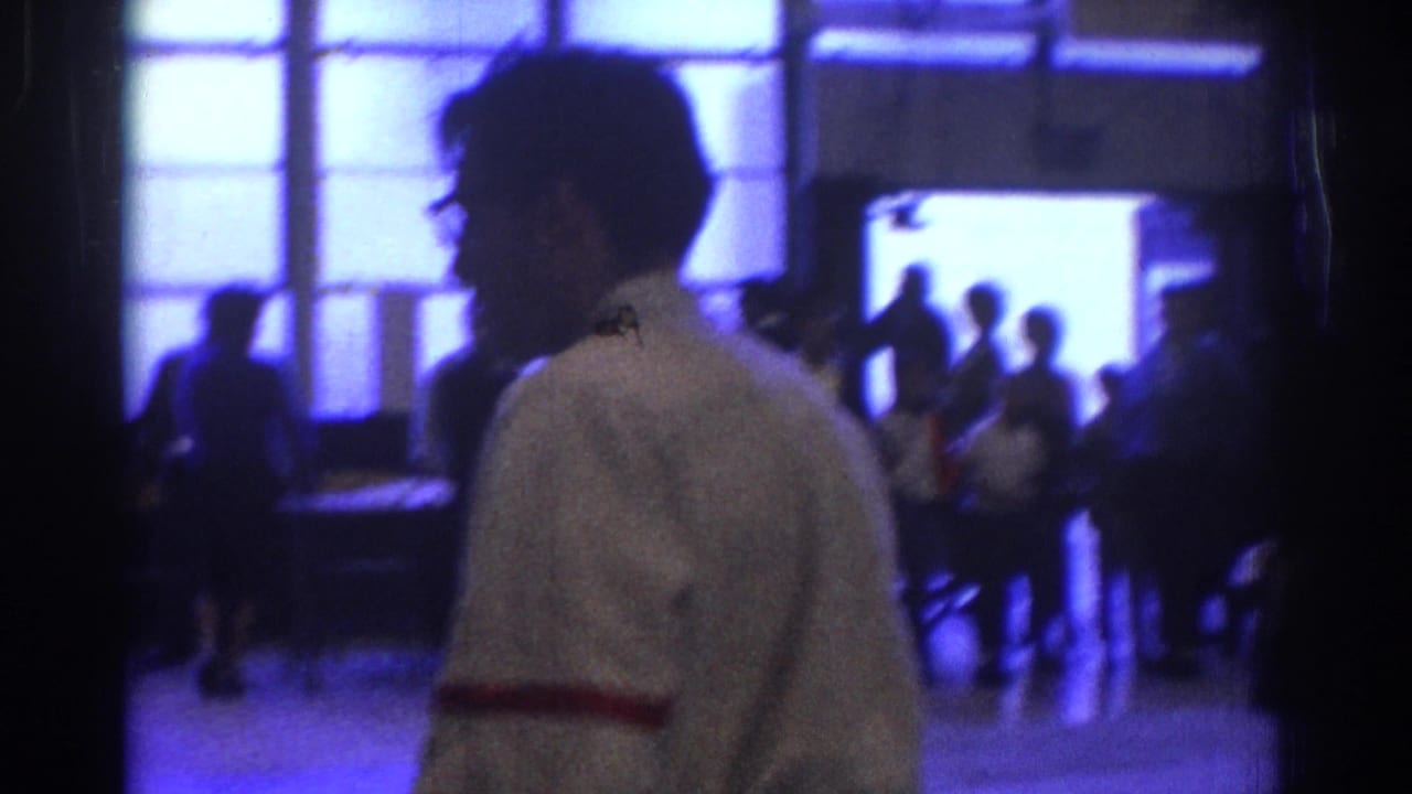 1966 West Covina home-movie frame with a white-shirted man in profile, blue-lit windows, and silhouettes of attendees in the background.
