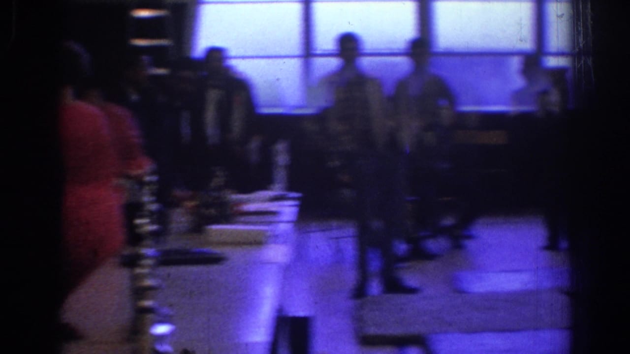 Dim hall with attendees near long tables and bright window panels.