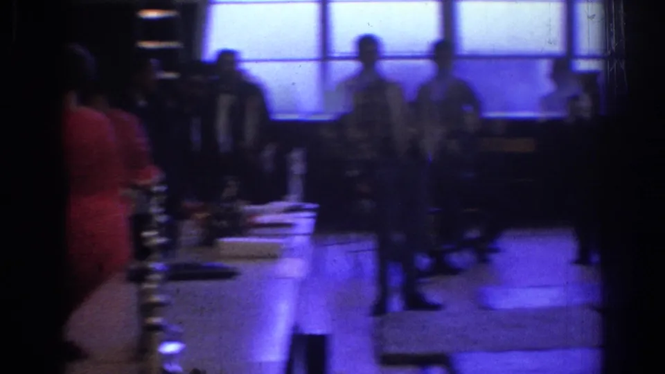 1966 West Covina 8mm frame showing a dim event hall with people standing near long tables and bright blue-tinted windows.