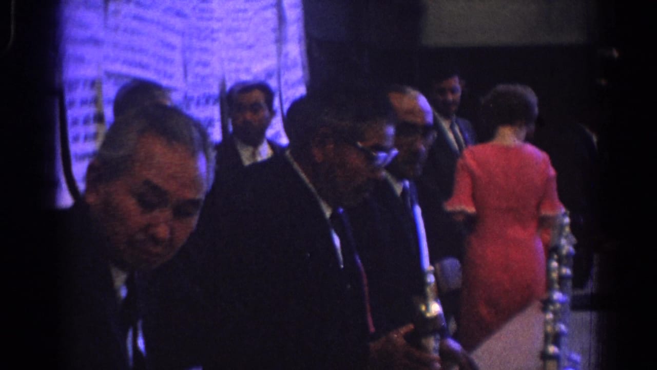 1966 West Covina frame with suited men and a woman in a red dress beside reflective award objects in a dim hall.