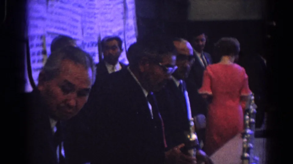 1966 West Covina frame with suited men and a woman in a red dress beside reflective award objects in a dim hall.