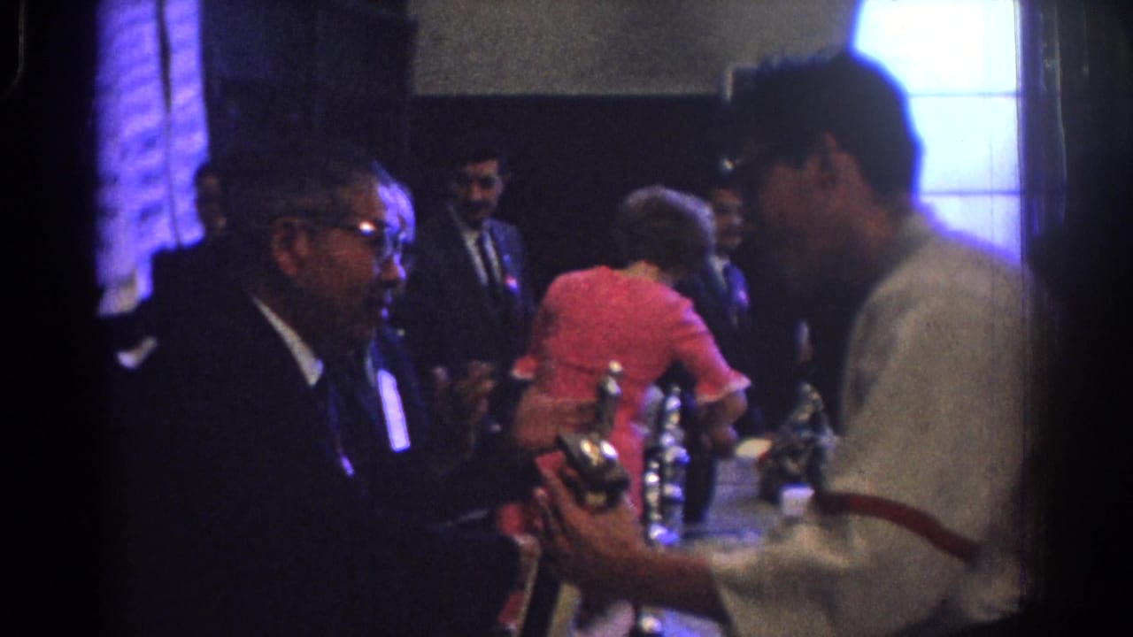 1966 West Covina frame showing a man in a light shirt holding a reflective award object near suited men and a woman in red.