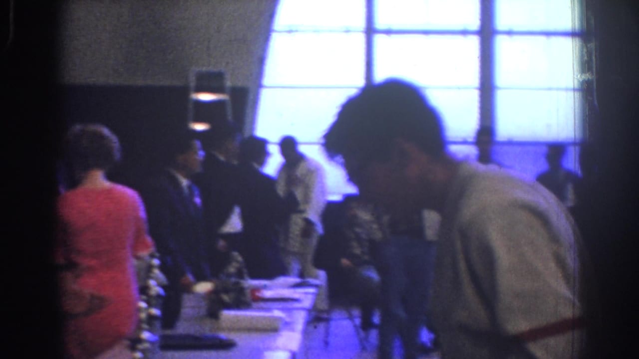 1966 West Covina 8mm frame with a man leaning over an awards table while attendees stand near bright window panels.