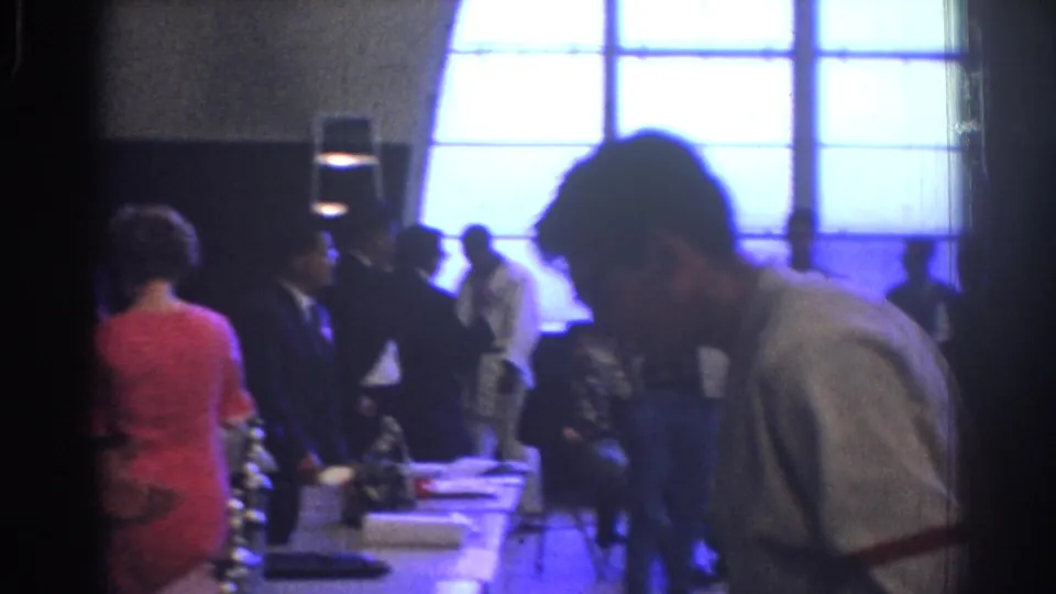 1966 West Covina 8mm frame with a man leaning over an awards table while attendees stand near bright window panels.
