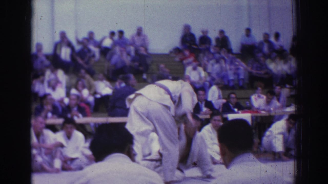 1966 West Covina judo competitors in white gi grapple on an indoor mat as spectators watch from bleachers.
