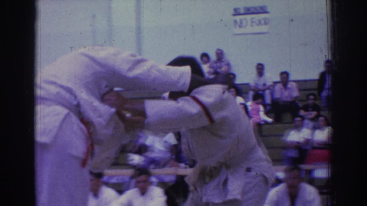 Close view of judo competitors in white gi during a forceful throw or hold, with an auditorium wall and spectators in the background.