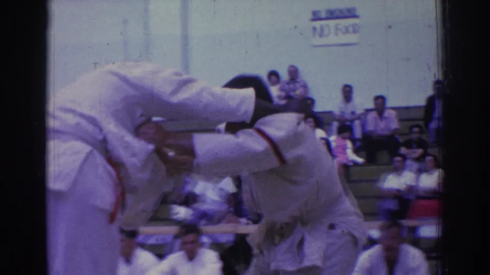 Close view of judo competitors in white gi during a forceful throw or hold, with an auditorium wall and spectators in the background.