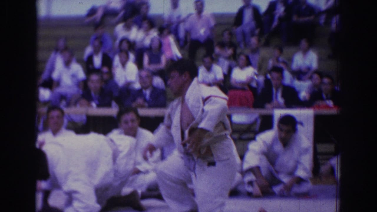 A judo athlete in a white gi stands near the mat after an exchange, with seated competitors and spectators in the auditorium behind him.