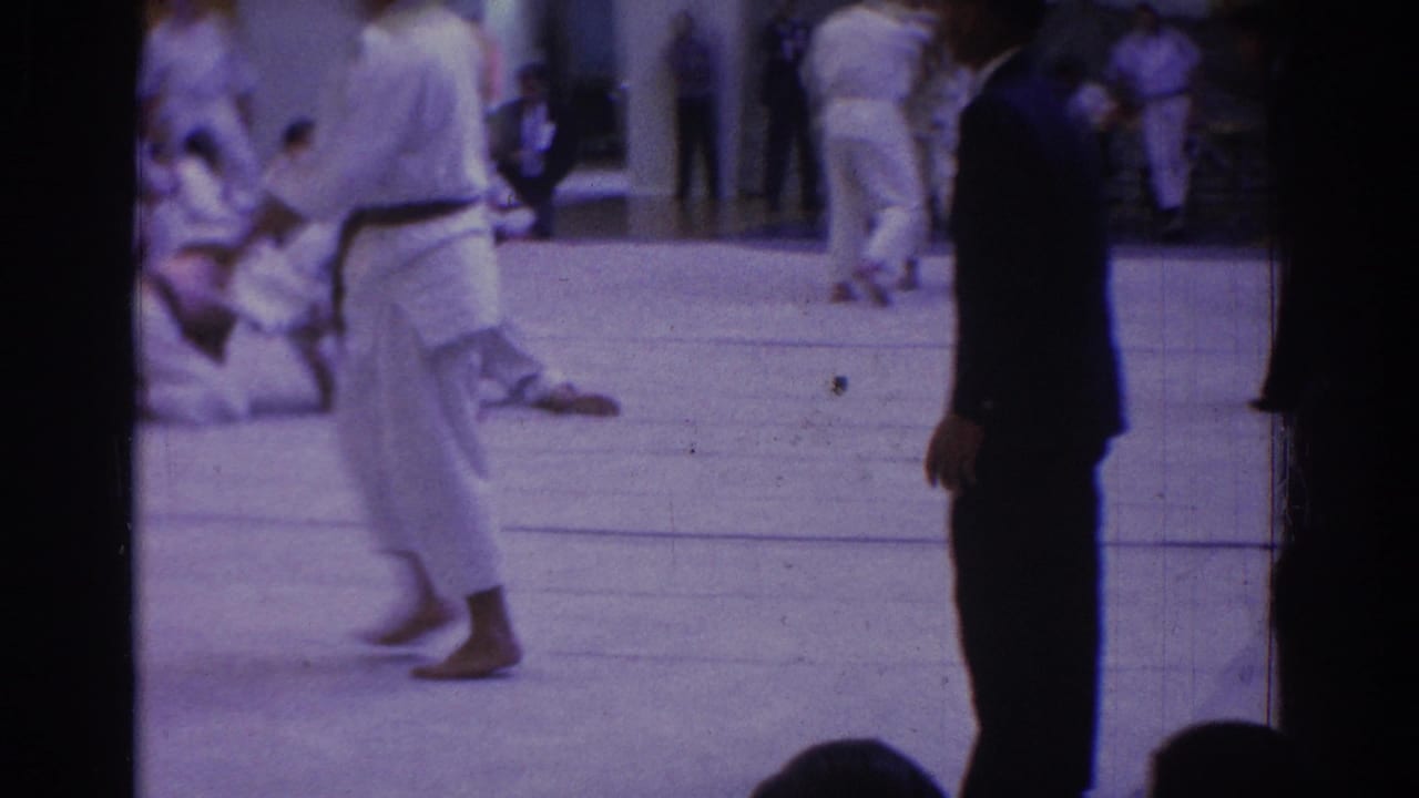 1966 West Covina judo competition footage showing white-uniformed competitors on a gym mat with a suited official or observer at the edge.
