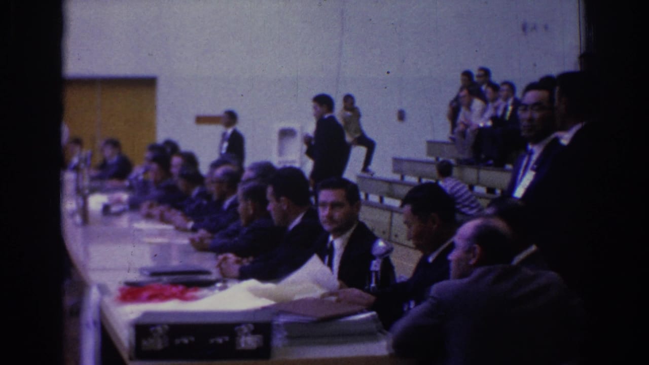 Officials sit at a long table beside gym bleachers during the judo competition.