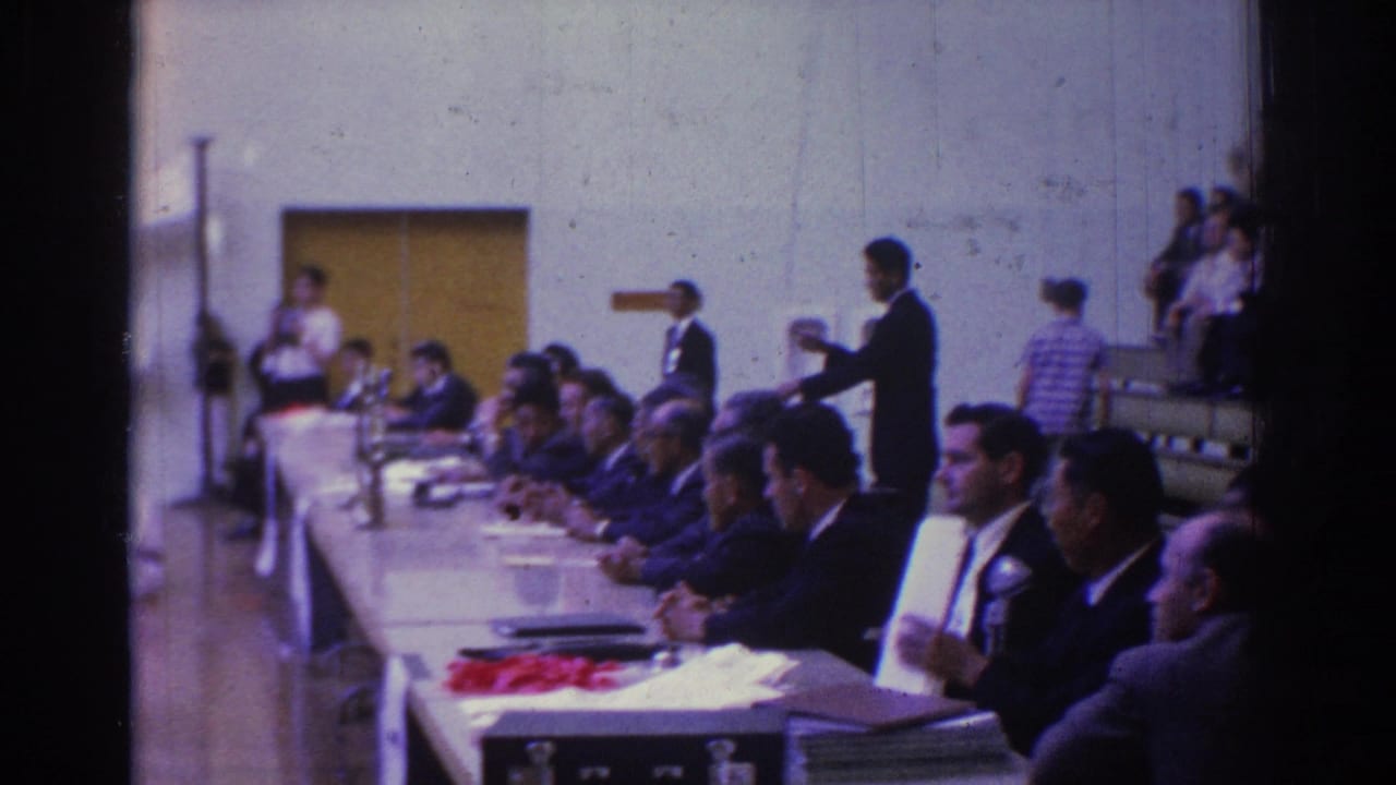 Wider 1966 gymnasium view with suited officials at a table, spectators on bleachers, and a standing figure near the wall.
