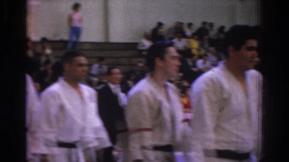 White-uniformed judo competitors move through the foreground while spectators sit on gym bleachers in the 1966 West Covina footage.