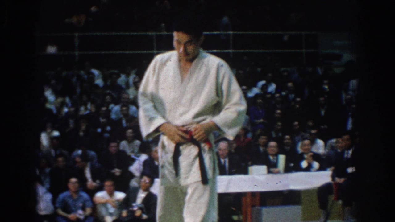 1966 West Covina tournament frame showing a karate competitor in white gi adjusting his black belt before spectators and officials.