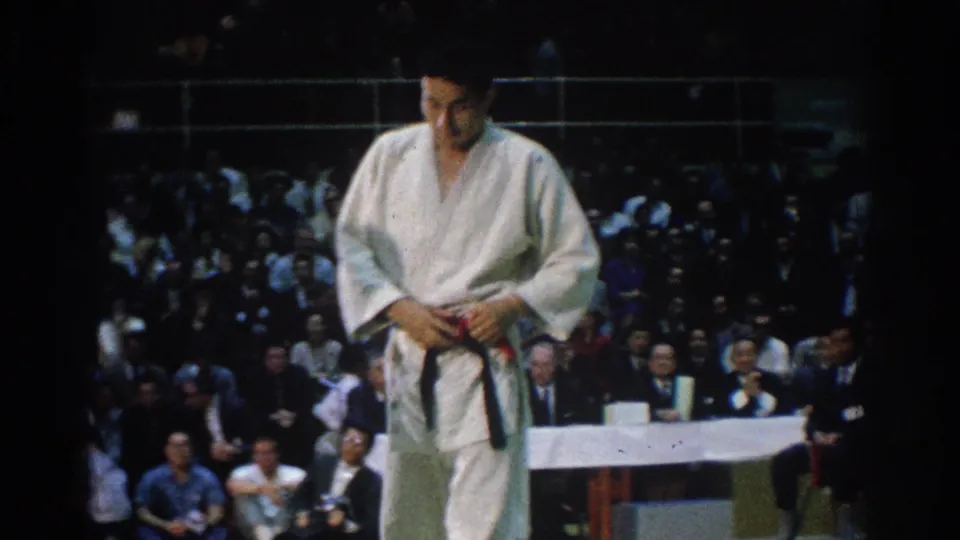 1966 West Covina tournament frame showing a karate competitor in white gi adjusting his black belt before spectators and officials.