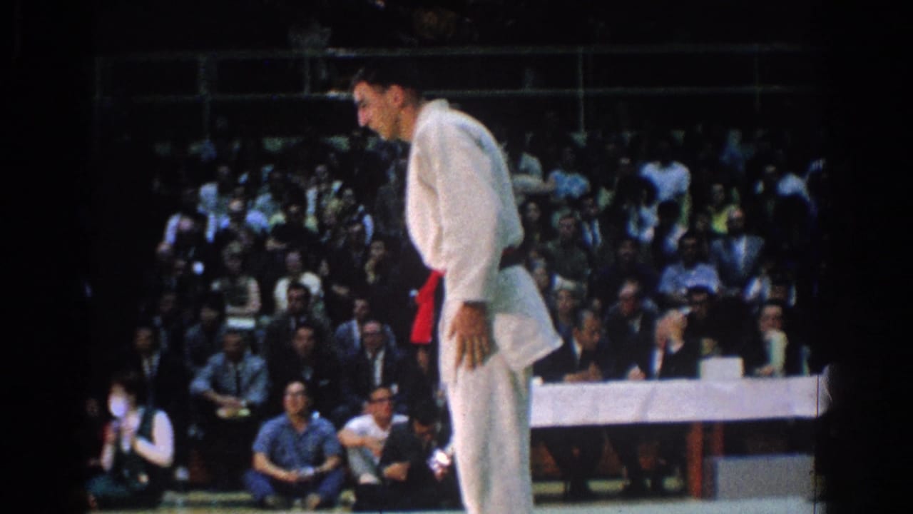 Judo athlete in a white gi stands on the mat before a seated audience.
