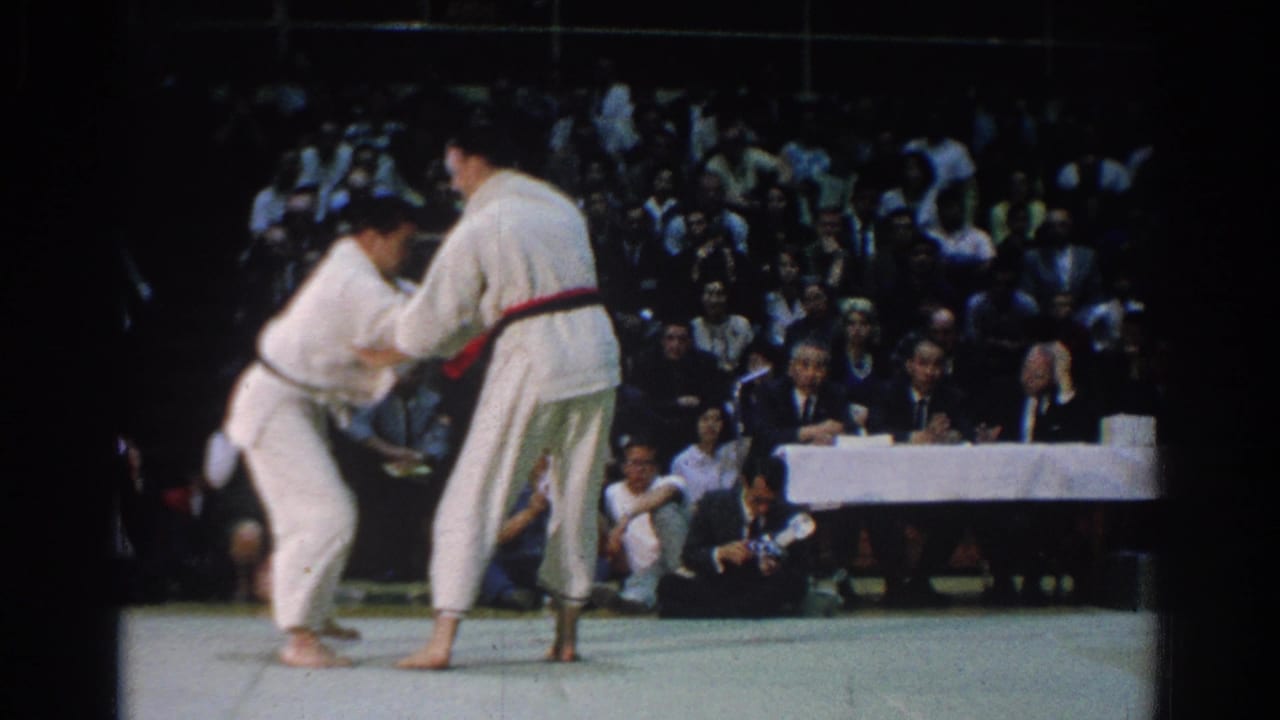 Two judo competitors in white gi grapple on a mat before seated officials and spectators.