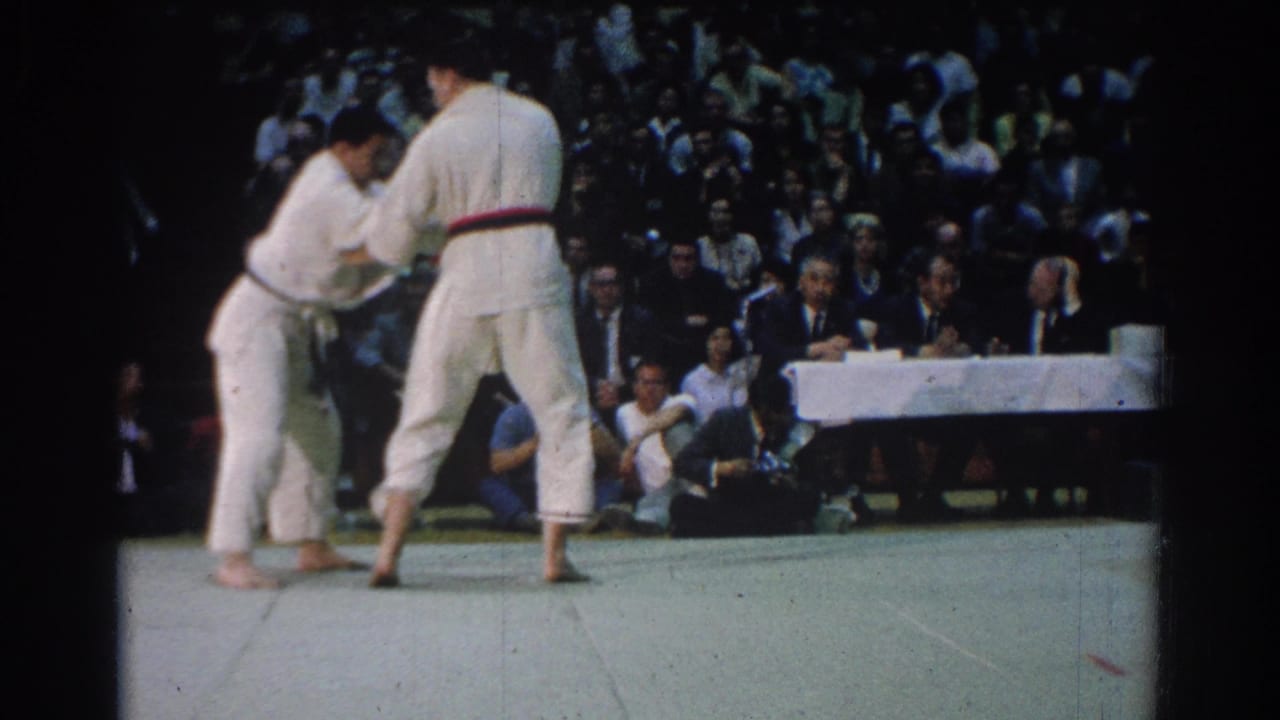 During the 1966 West Covina match, two male competitors in white gi continue gripping while spectators fill the background.