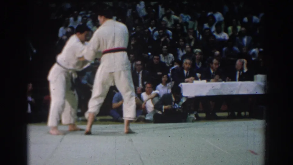 During the 1966 West Covina match, two male competitors in white gi continue gripping while spectators fill the background.