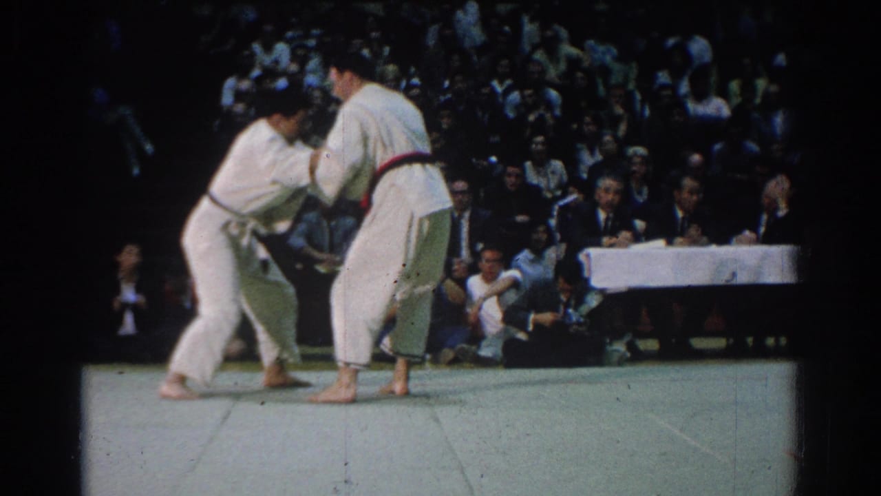 A close judo grip unfolds on the mat in West Covina, with judges seated at a long table and spectators behind them.