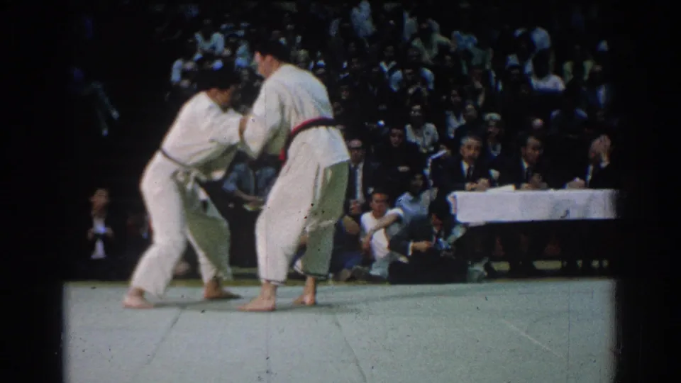 A close judo grip unfolds on the mat in West Covina, with judges seated at a long table and spectators behind them.