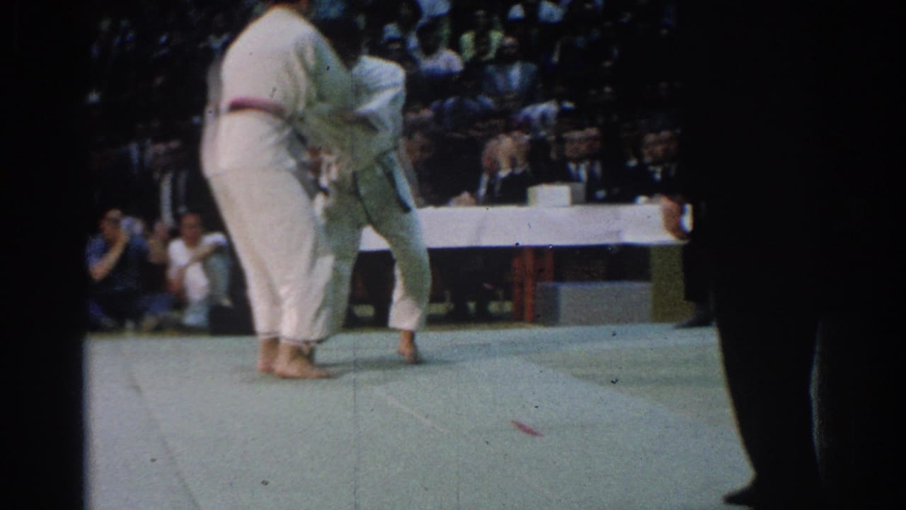 1966 West Covina footage showing two male martial arts competitors in white gis grappling on a green mat before a seated audience.