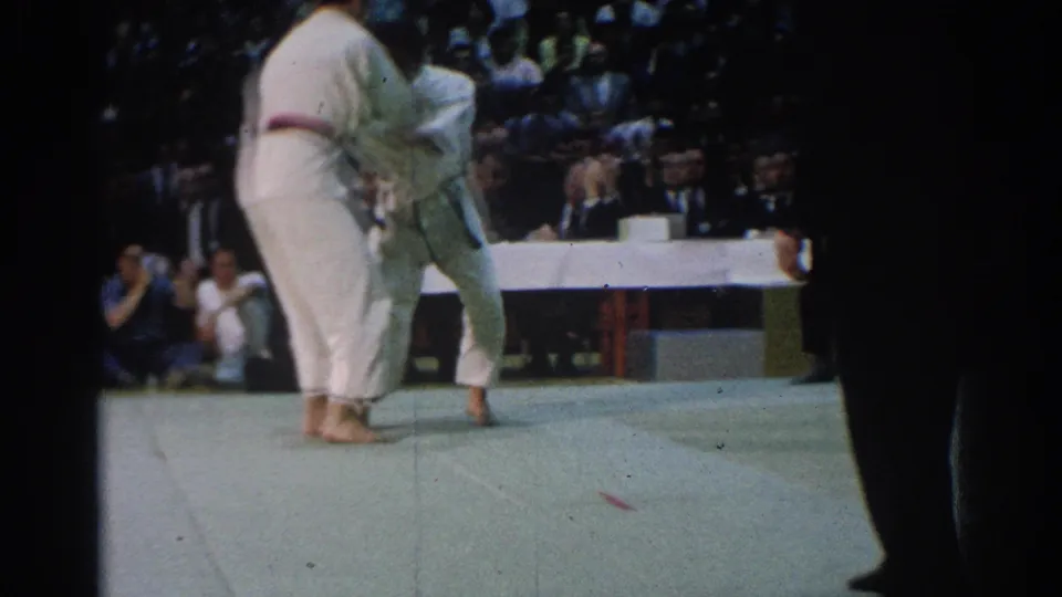 1966 West Covina footage showing two male martial arts competitors in white gis grappling on a green mat before a seated audience.