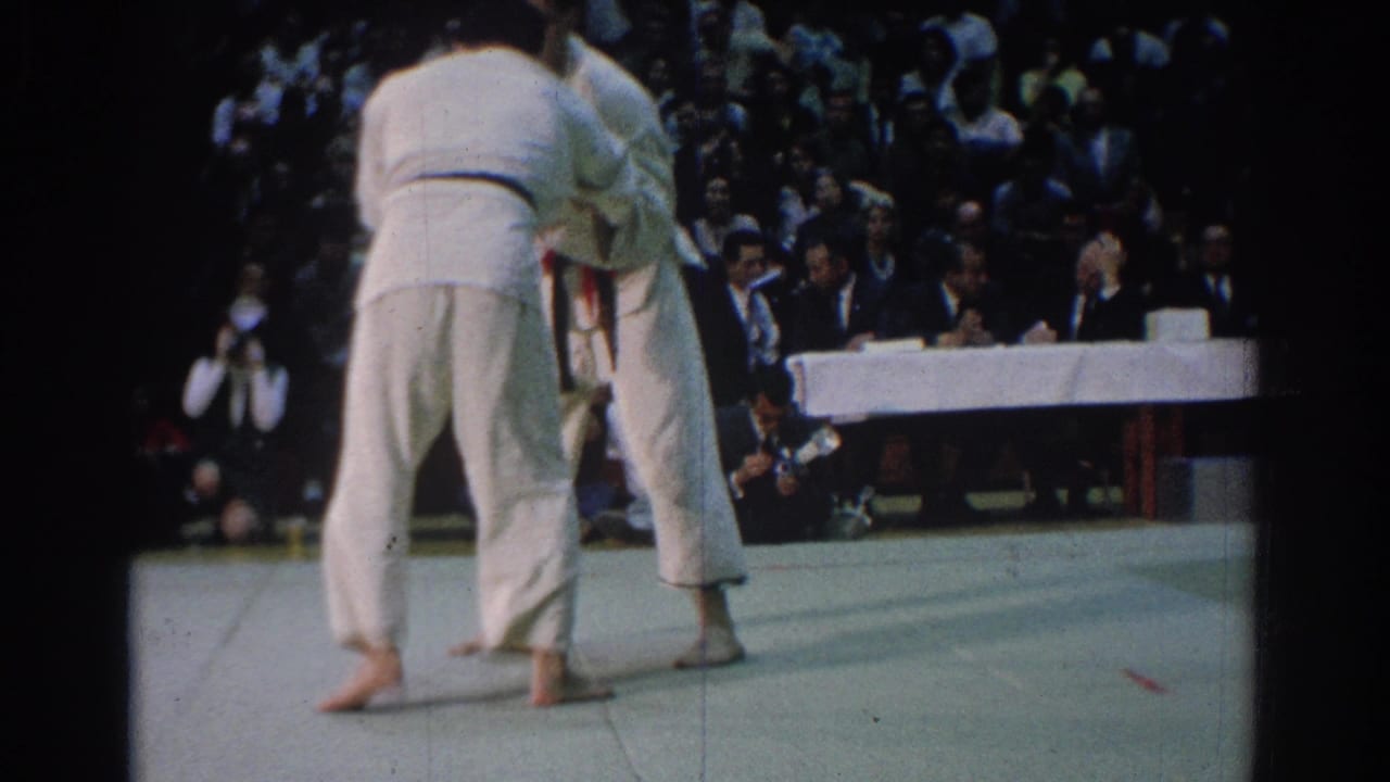 Two judokas in white gi grapple on a mat before a seated crowd.