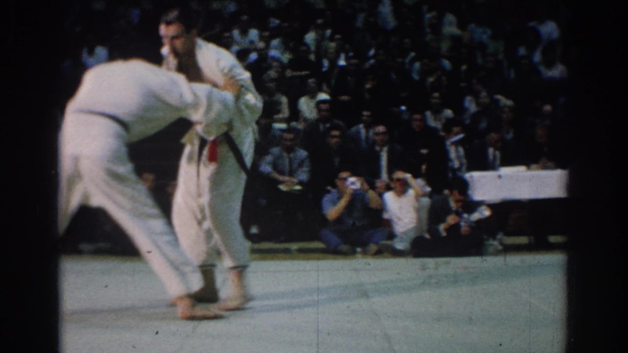 A 1966 West Covina judo bout continues as one competitor leans into a grip while spectators and photographers watch.