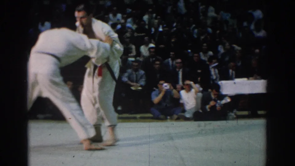 A 1966 West Covina judo bout continues as one competitor leans into a grip while spectators and photographers watch.