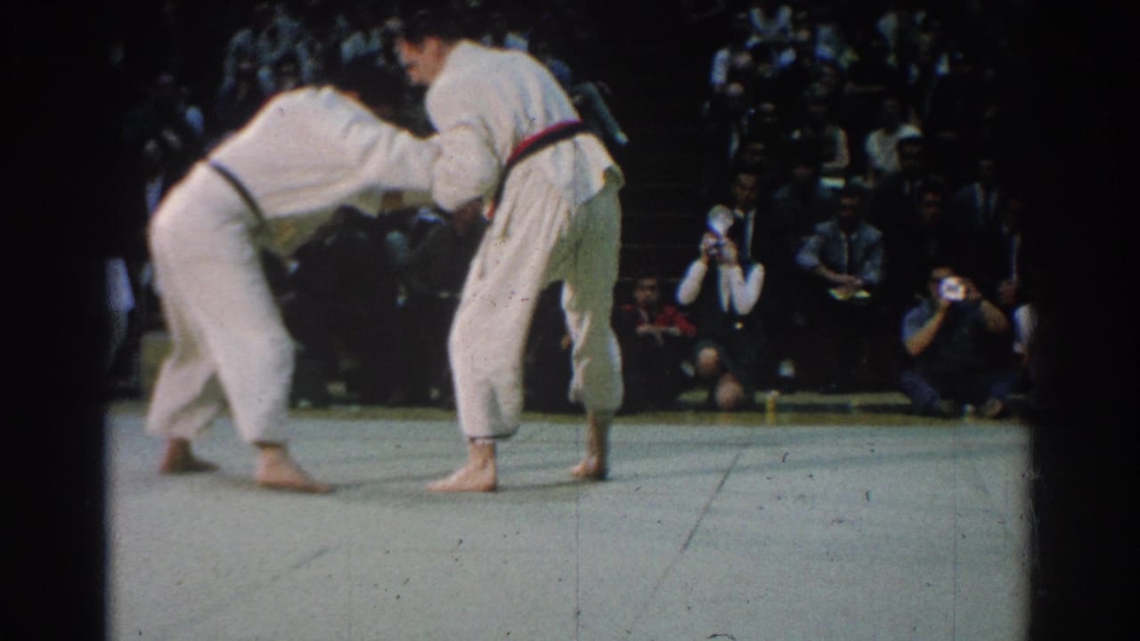 Two barefoot judokas in white uniforms bend low in a standing grip during the West Covina match, with cameras visible behind them.