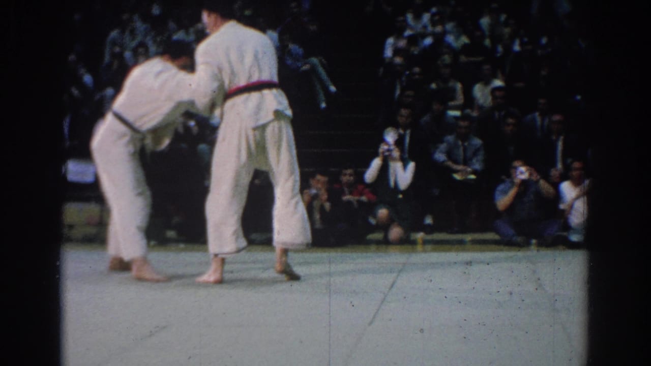 Near the end of the clip, the 1966 West Covina judo competitors remain upright in a grip before a dense seated audience.