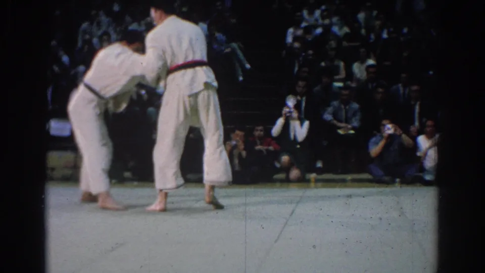 Near the end of the clip, the 1966 West Covina judo competitors remain upright in a grip before a dense seated audience.
