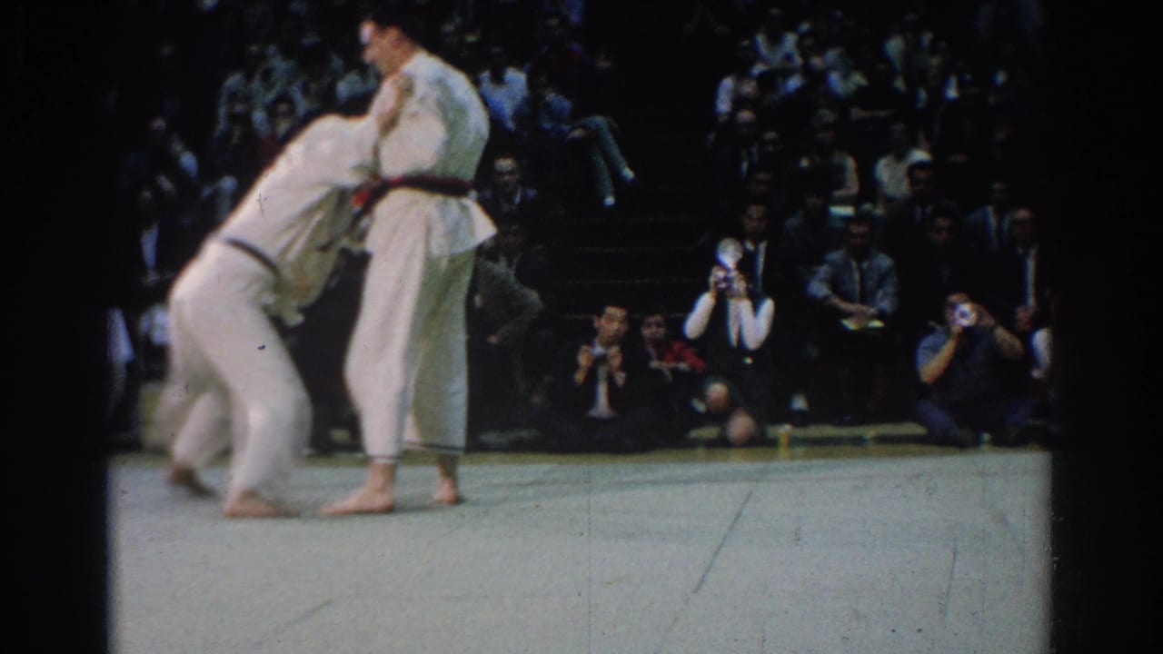 Two judo competitors in white uniforms grapple on a mat before a seated audience.