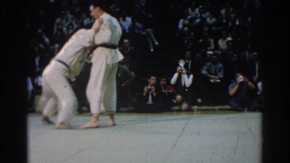 In West Covina, California in 1966, two judo competitors in white gi uniforms grapple on a pale mat as spectators watch from the background.