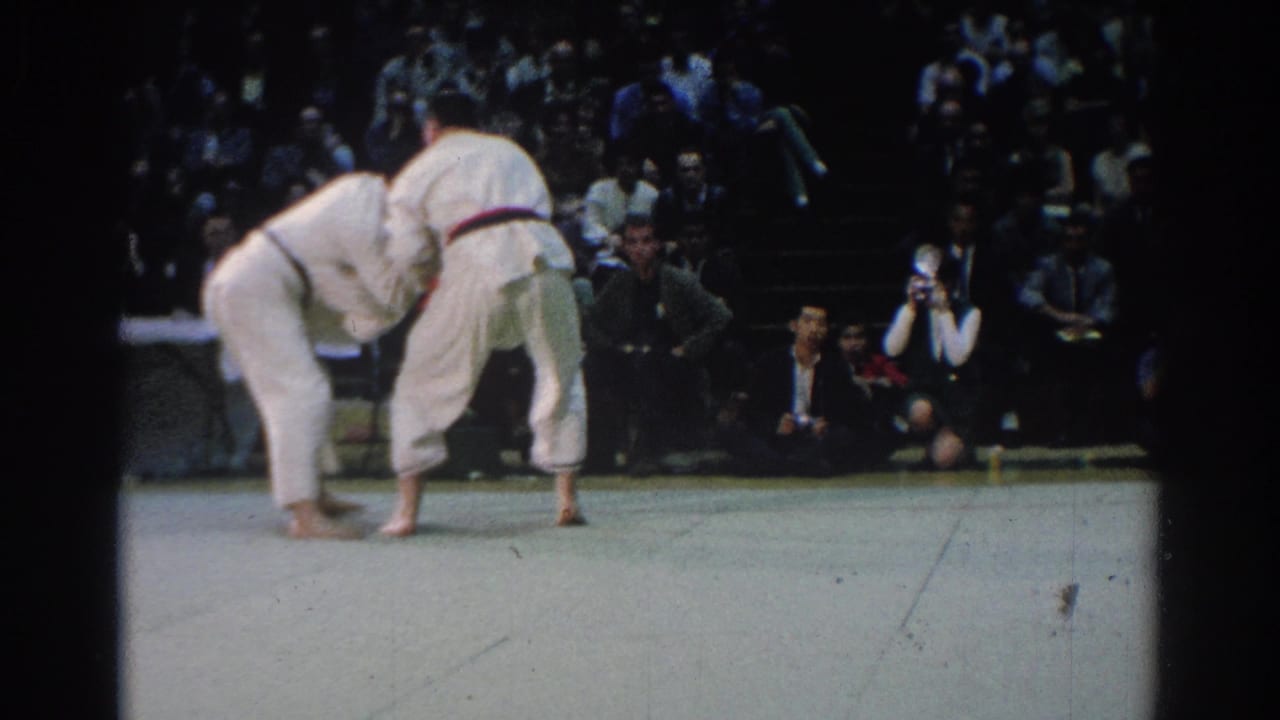 Two judo competitors in white uniforms lean into a throw or off-balance movement during the 1966 West Covina match, with a crowd seated behind them.