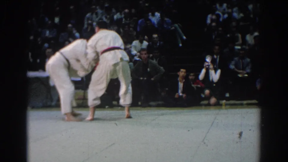 Two judo competitors in white uniforms lean into a throw or off-balance movement during the 1966 West Covina match, with a crowd seated behind them.