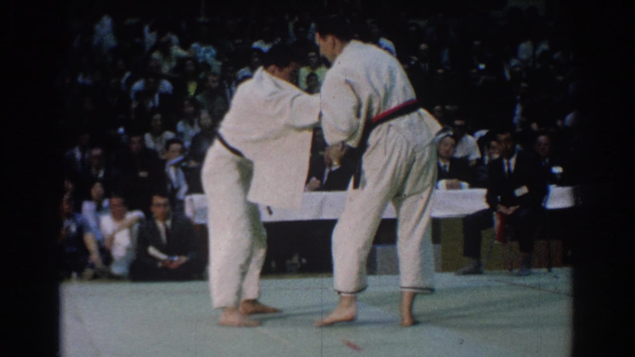 During the West Covina judo match, the fighters stand upright in a clinch on the mat while a crowded gym audience watches closely.